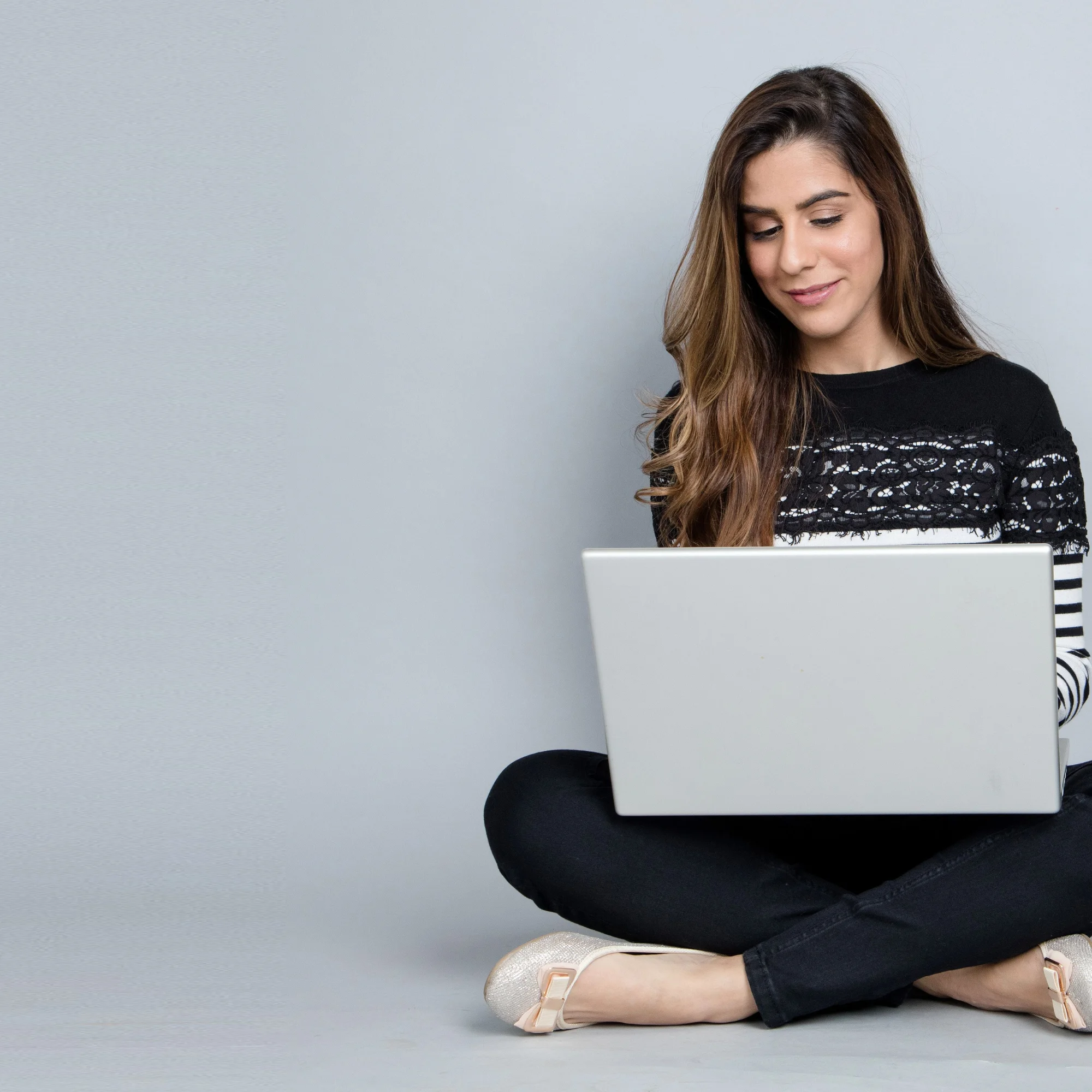 A young woman sitting cross-legged on the floor, smiling while working on a laptop, wearing a black and white striped top with black pants, set against a minimal grey background